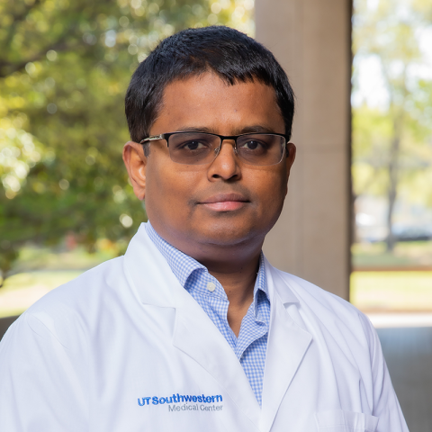 a man with dark short hair and glasses wearing a white lab coat in an outside environment