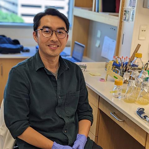 A man sits by a lab bench smiling with a dark grey-blue shirt and lab gloves