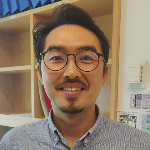 Smiling man with thick dark hair, mustache, and goatee, wearing a blue checked shirt and dark-rimmed glasses.