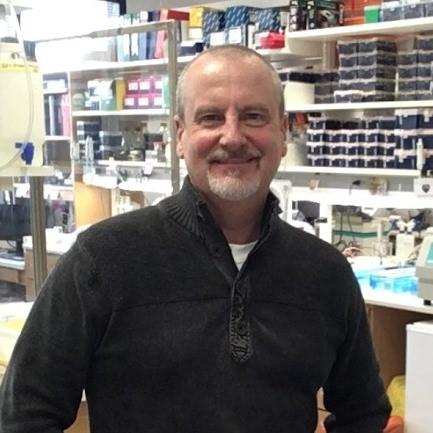 Dr. Farrer, smiling man with close-cropped hair, beard and mustache, wearing a black shirt, standing in a lab.