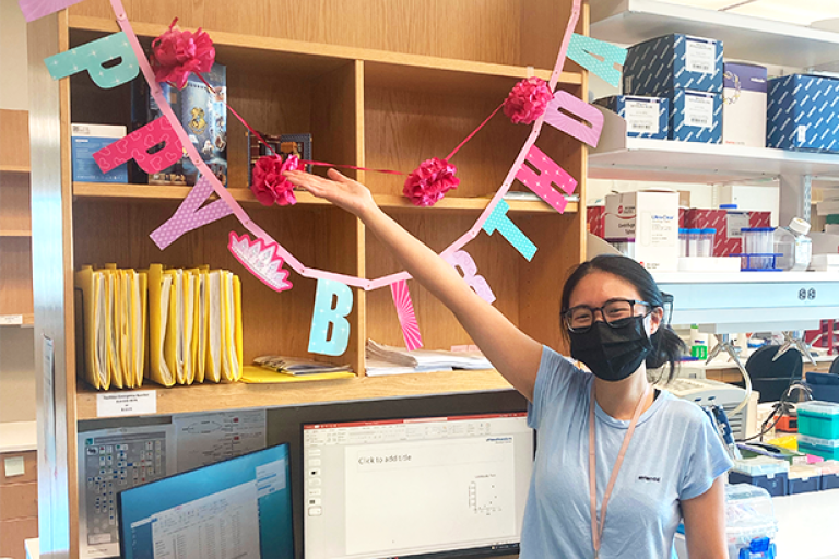 Maggie wearing face mask, standing with "Happy Birthday" banner