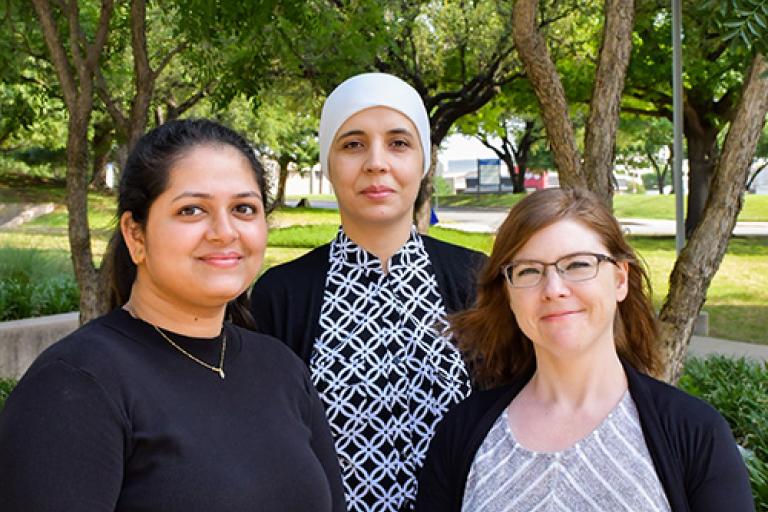 Three smiling women standing in front of a grove of trees.