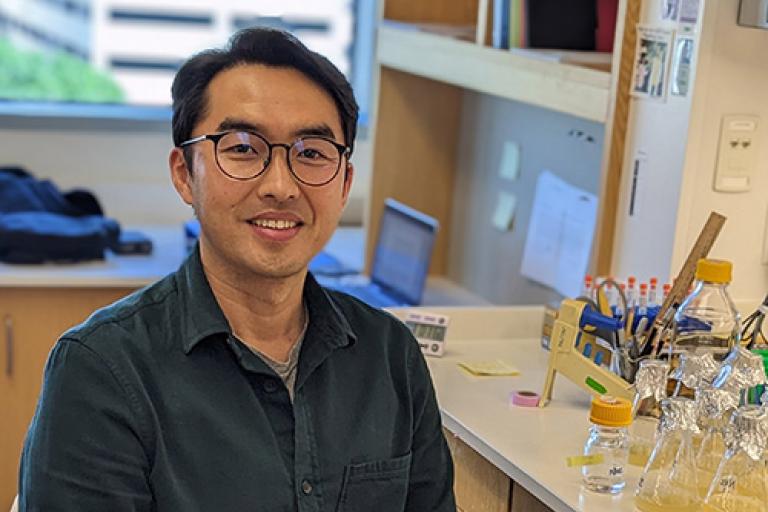 A man sits by a lab bench smiling with a dark grey-blue shirt and lab gloves