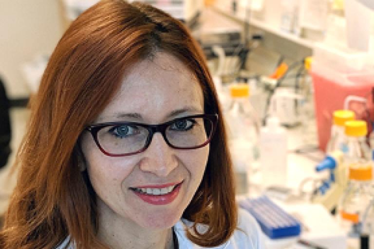 A woman with red hair and glasses sits by a lab bench smiling