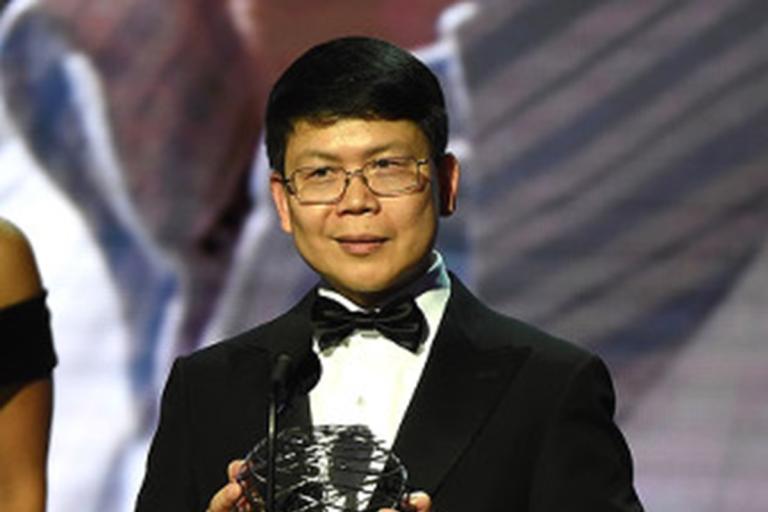 Smiling man with dark hair and glasses, wearing a tuxedo, holding a globe-shaped award.
