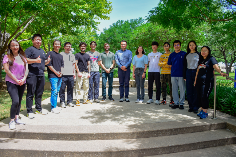 Group of 12 lab members outside with large trees in the background.