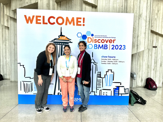 Alex, Maggie, and Anju standing and smiling in front of a poster decorated with the Seattle skyline