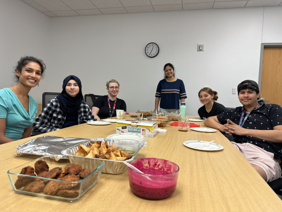 Anju, Zehra, Timea, Maggie, Alex, and Abner seated at a table covered with a variety of foods.