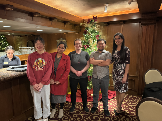 Bingbing, Maggie, Timea, Sebastian, and Anju standing in front of a decorated tree and smiling.