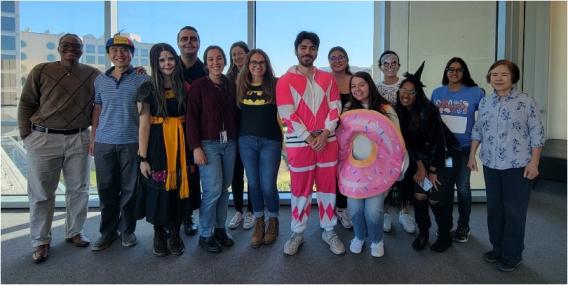 14 lab team members pose in front of a large window, some in costumes.