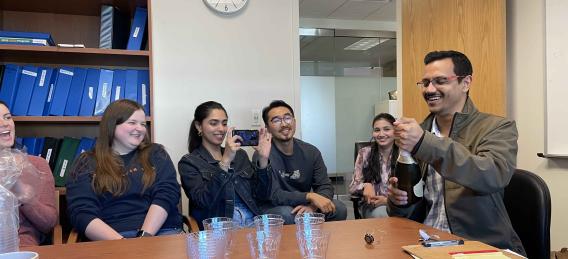 One man opens a bottle and four women and one man look on smiling and laughing.