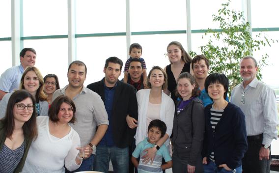 The Farrer Lab team and some family members posing in front of a large window and a green plant.