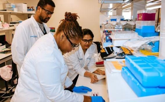 3 researchers in lab wearing white coats, safety glasses, and blue surgical gloves.