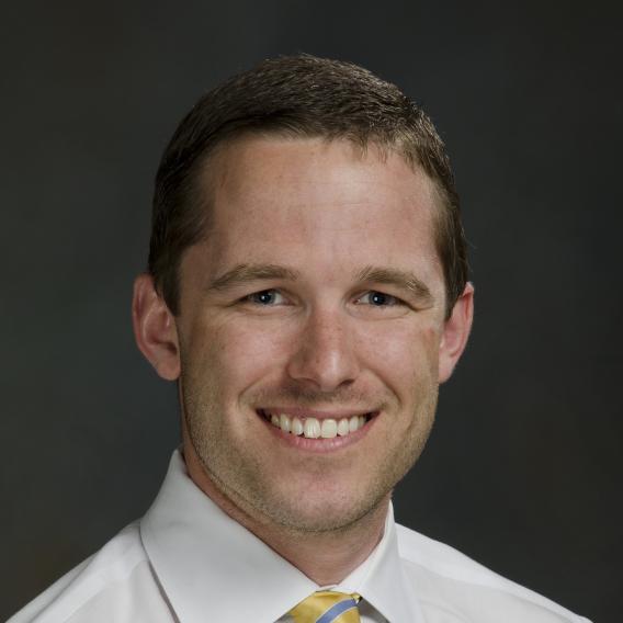 Smiling man with light brown hair, wearing a white dress shirt with a gold tie with stripes in two shades of blue.