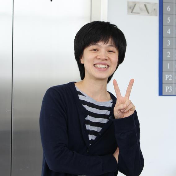 Smiling woman with short dark hair, wearing a dark sweater over a striped shirt, holding up two fingers in a "Peace" sign.