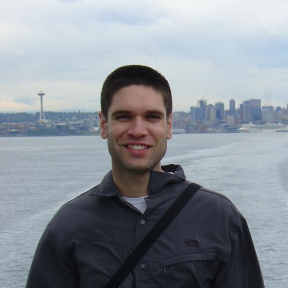 Dr. Huber, smiling, wearing a dark jacket, outdoors with water and a city in the background.