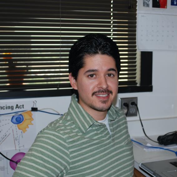 Dr. Ramos, with dark hair and a trim beard and mustache, wearing a green striped shirt, sitting in front of a window covered with mini-blinds.