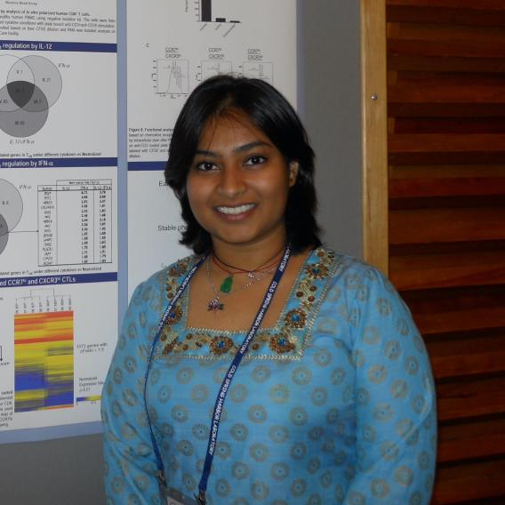 Dr. Chowdhury, smiling, with shoulder-length dark hair, wearing a blue print dress, standing in front of a a bulletin board.