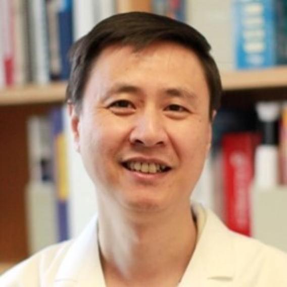Gang Huang wearing lab coat, standing in front of shelves of books.