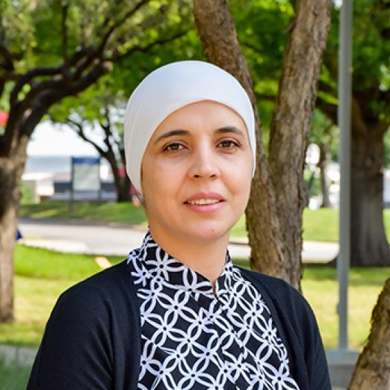 Smiling woman wearing a head scarf and blue sweater standing outside among trees.