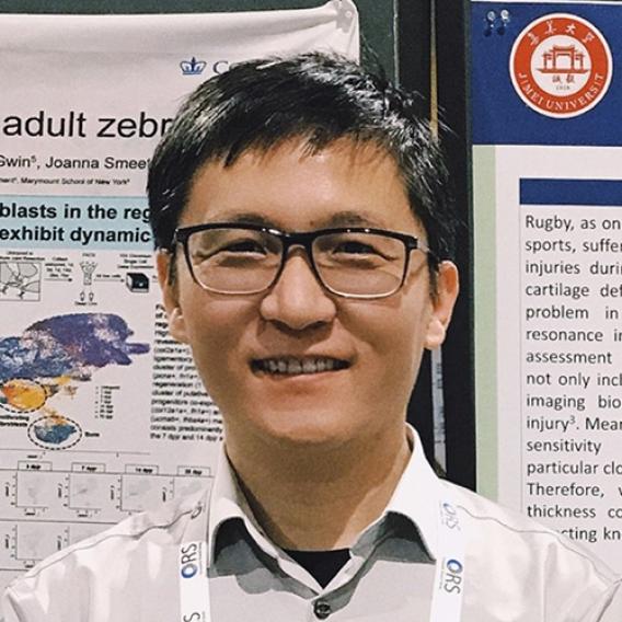 Smiling man with dark hair and glasses, standing in front of a bulletin board.