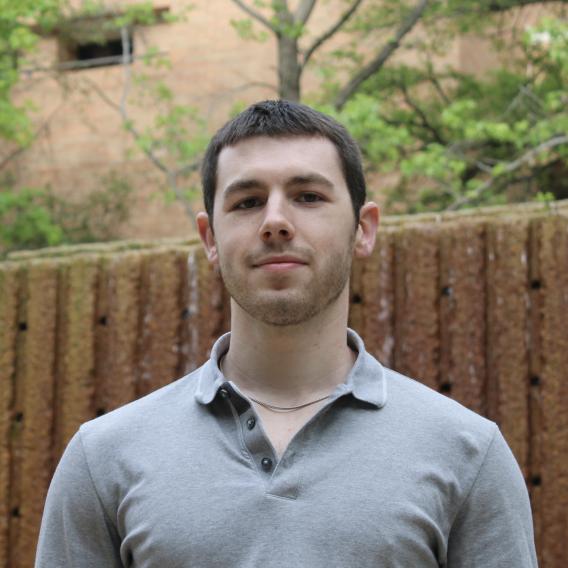 Man with brown hair, outdoors, wearing a gray shirt.