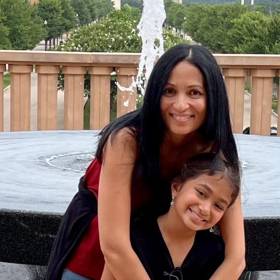 Smiling woman with long black hair sitting next to a fountain and railing, with her arms around a little girl.