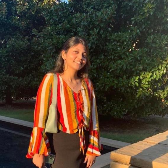 Smiling woman with dark hair, outside next to a bush, wearing a blouse with orange, white, and yellow vertical stripes.