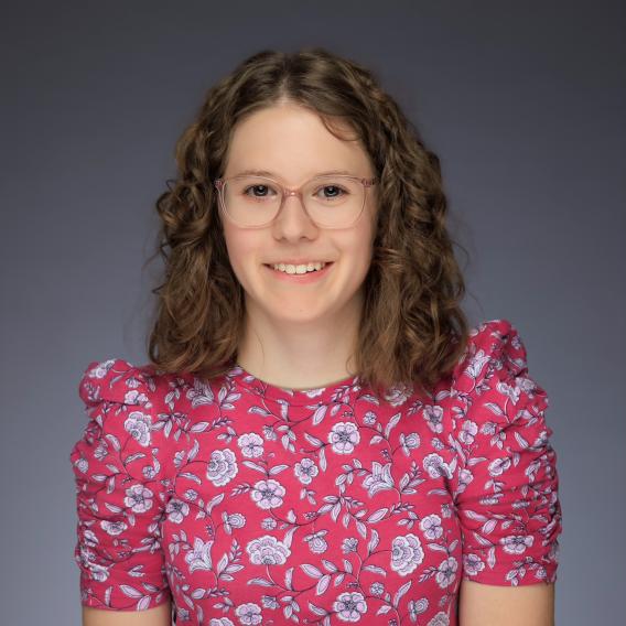 Smiling woman with dark hair, outside next to a bush, wearing a blouse with orange, white, and yellow vertical stripes.