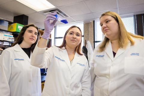 Emily Nettesheim, Katherine Wert, Ph.D., and Ashley Rowe in white lab coats looking at a test tube Dr. Wert is holding up.
