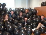 Six team members in office buried up to their necks in black balloons