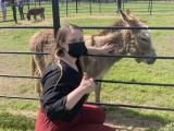 Woman wearing a pandemic mask, petting a donkey and giving a thumbs-up at an outdoor paddock
