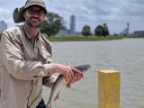 Mike holds a channel catfish. Behind him is the Trinity River, and the Dallas skyline is visible in the distance