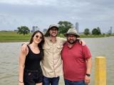 From left to right: Anna, Mike, and Marco pose for a photo in front of the Trinity River. The Dallas skyline is visible behind them. 