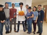 8 lab members posed in a hallway, some wearing hats. Man in the middle is holding a jack-o-lantern.