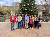 9 lab members posed outside in front of a large Christmas tree, most wearing holiday sweaters.