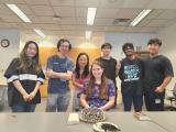 Dr. Dan Tong with lab members gathered around a woman sitting in front of a cake