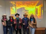 Five individuals in cowboy hats and western attire in front of a western movie style image. 
