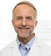 Smiling man with short fair hair, beard, and mustache, wearing a white lab coat over a blue shirt.