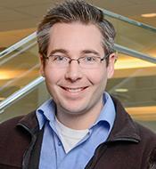 Smiling man with short, curly salt-n-pepper hair, wearing a brown jacket over a blue shirt with glasses.