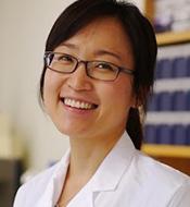 Smiling woman with black hair, wearing a white lab coat and dark rimmed glasses.