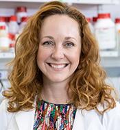 Smiling woman with long curly red hair, wearing a white lab coat over a print blouse.