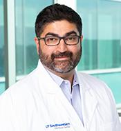 Man with thick black hair, mustache, and black and gray beard, wearing black-rimmed glasses and a white lab coat.