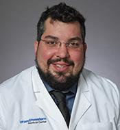 Smiling man with curly dark hair, beard, and mustache, wearing a white lab coat and glasses