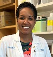 Smiling woman with short black curly hair, wearing a white lab coat over a floral print top.