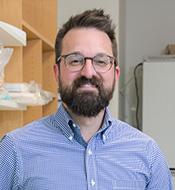 Smiling man with spiky brown hair, full beard, and mustache, wearing a blue shirt and glasses.