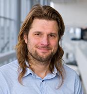 Smiling man with long brown hair, thin beard and mustache, wearing a blue shirt.