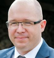 Smiling man with shaved head, wearing a blue suit, white shirt, and glasses.