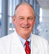 Smiling man with receding gray hair, wearing a white lab coat over a blue shirt and red plaid tie.