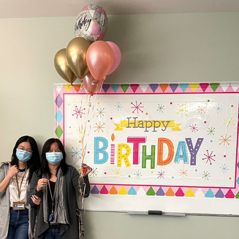 Kelly and Wei wearing masks, holding balloons, and standing in front of "Happy Birthday" banner